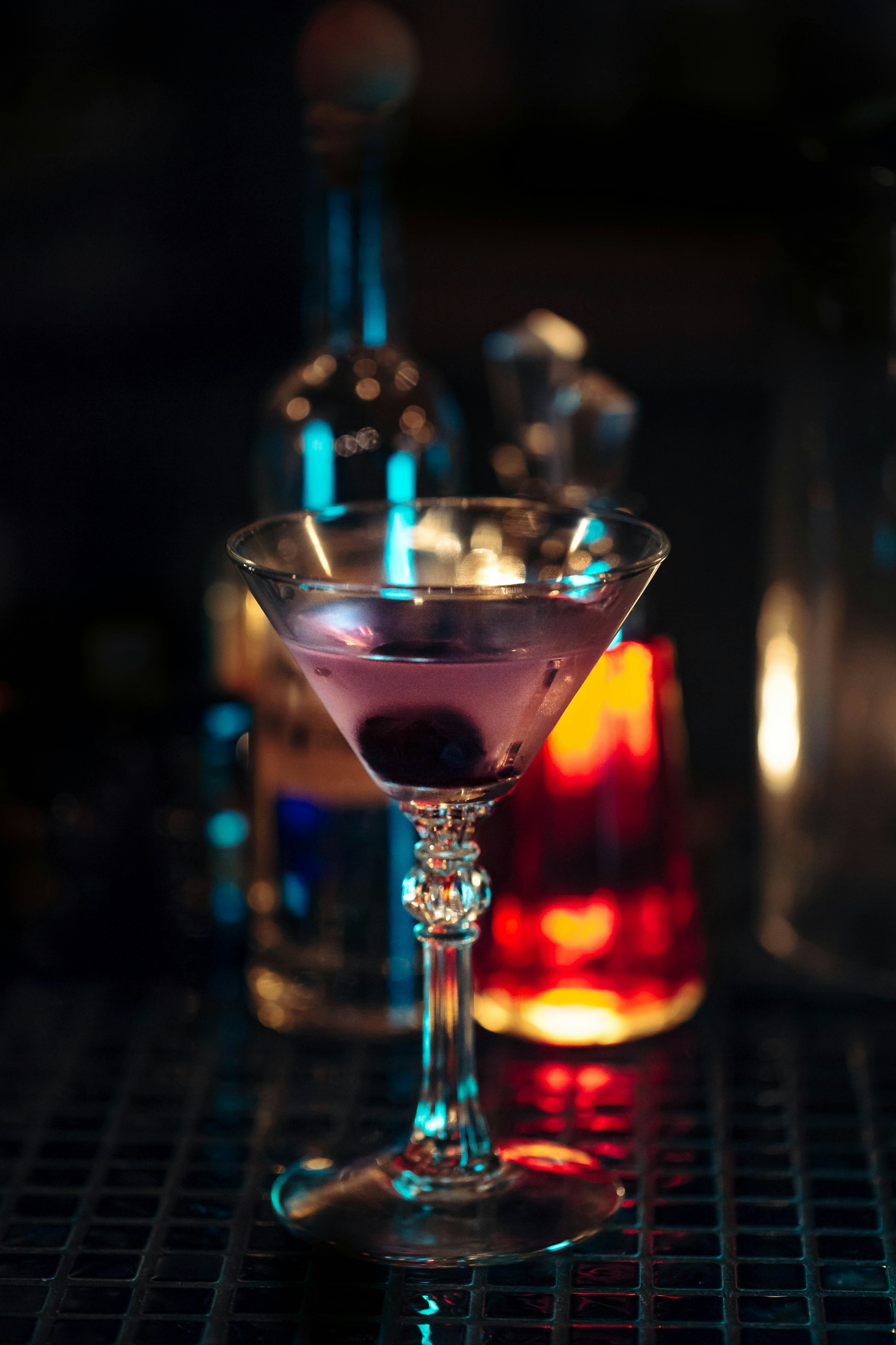 A coupe cocktail on the bar, bottles softly out of focus in the background under low light